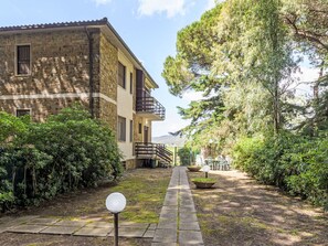 Edifici de pedra amb un balcó, envoltat de vegetació i un camí que condueix a una zona d'estar.