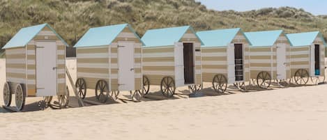 A row of beach huts with a simple design, each with a door and a small window, situated on a sandy beach with dunes in the background.
