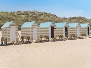 En række strandhuse med et enkelt design, hvert med en dør og et lille vindue, beliggende på en sandstrand med klitter i baggrunden.