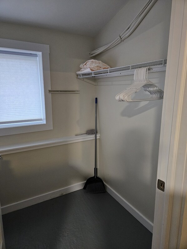 Een kleine, smalle kamer met een raam, een bezem, een dweil en een kledingrek.