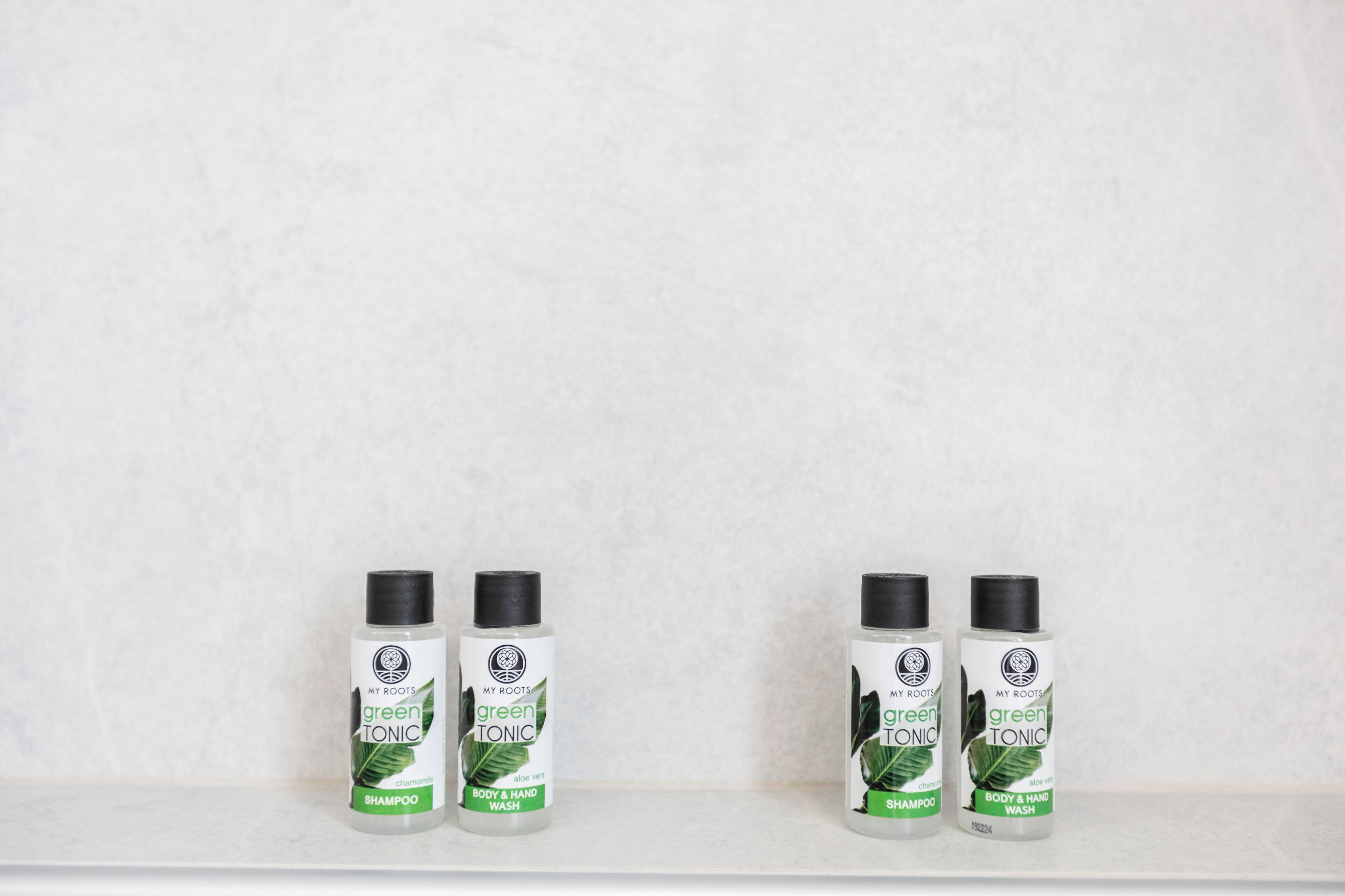 Fire flasker med grøn shampoo og shower gel på en hvid hylde foran en ensfarvet væg.