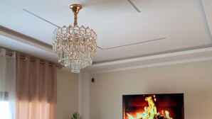 A living room with a chandelier, a flat-screen TV displaying a fire, a sofa, and a potted plant. - Villa sabrina gold (Djerba Midun)