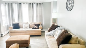 Una sala de estar con un sofá color beige, dos otomanas, un reloj en la pared y cortinas traslúcidas.