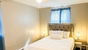 Un dormitorio con cabecera capitonada, cama con ropa de cama blanca, dos mesitas de noche con lámparas y una ventana con cortinas.