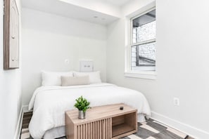 Ein Schlafzimmer mit einem Bett, einem Nachttisch mit Pflanze, einem Fenster und einer Tür.