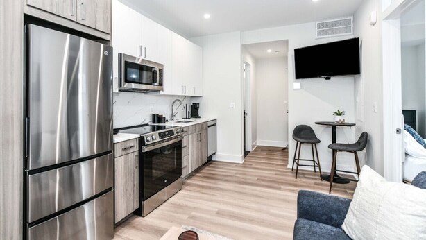 A modern kitchen with stainless steel appliances, a dining area with a small table and two chairs, and a flat-screen TV mounted on the wall.