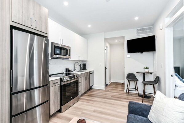 A modern kitchen with stainless steel appliances, a dining area with a small table and two chairs, and a flat-screen TV mounted on the wall.