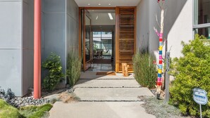 Ingresso moderno di un edificio con porta in vetro, una panchina in legno e un vialetto fiancheggiato da piante.