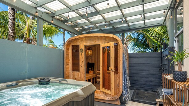 Una vasca idromassaggio e una struttura sauna in legno con porta in vetro, circondate da un pontile in legno e palme.
