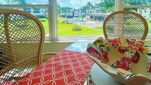Una habitación con dos sillones de mimbre, una mesa con mantel estampado, y vistas a una piscina y edificios a través de amplios ventanales.
