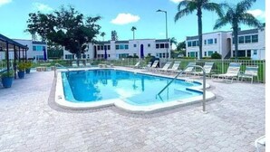 Una piscina con sillas de descanso, un área de estar techada y un edificio con varias ventanas.