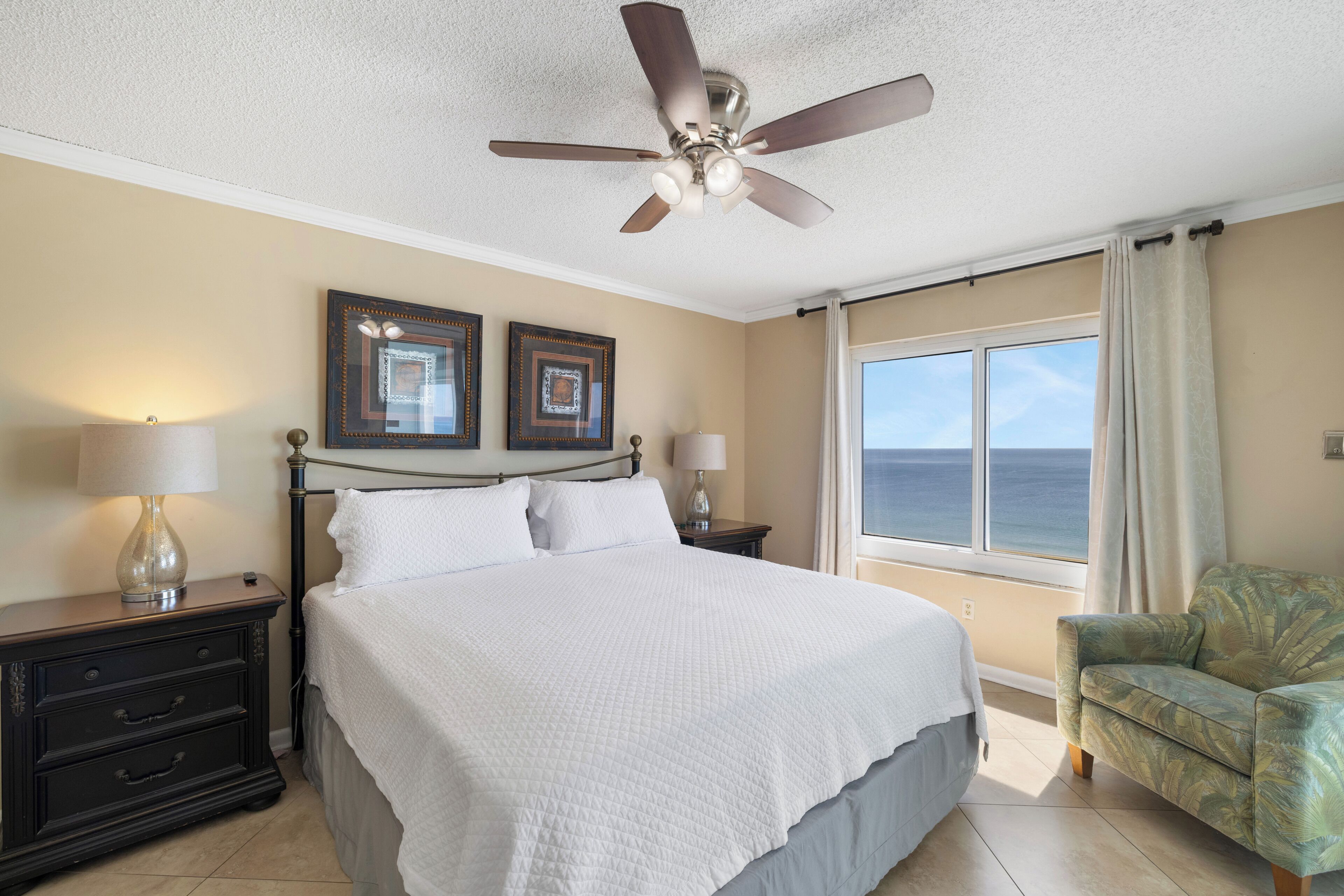 A bedroom with a bed, two nightstands, a ceiling fan, a window with a view, and a chair.