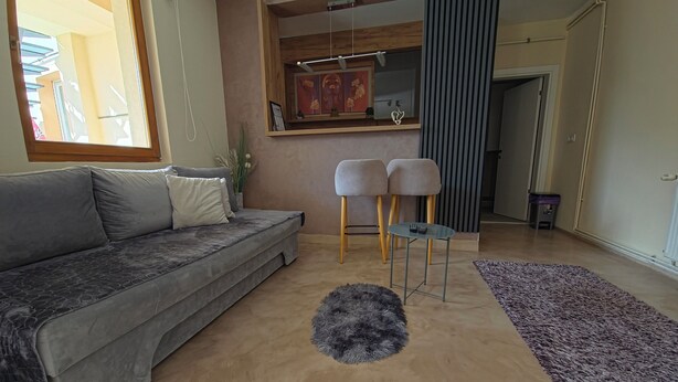Ein modernes Wohnzimmer mit einem grauen Sofa, zwei hohen Barhocker, einem Glastisch, einem flauschigen Teppich und einem großen Fenster.