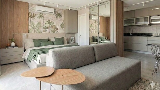 Um quarto moderno com sofá, mesas de cabeceira, espelho e uma pequena cozinha.