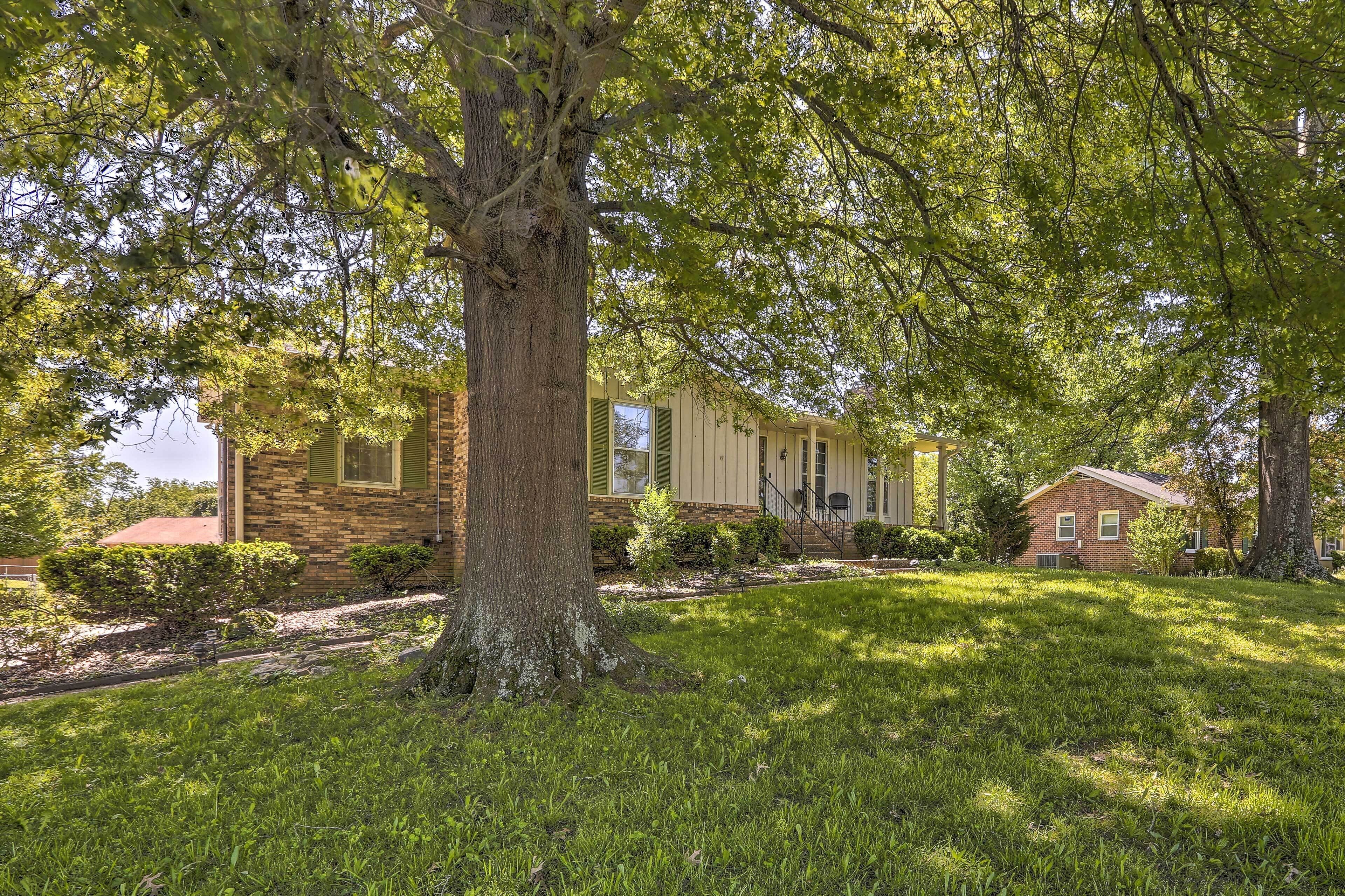 A residential property with a large tree in the front yard, a brick house with a red roof, and a smaller brick building in the background.