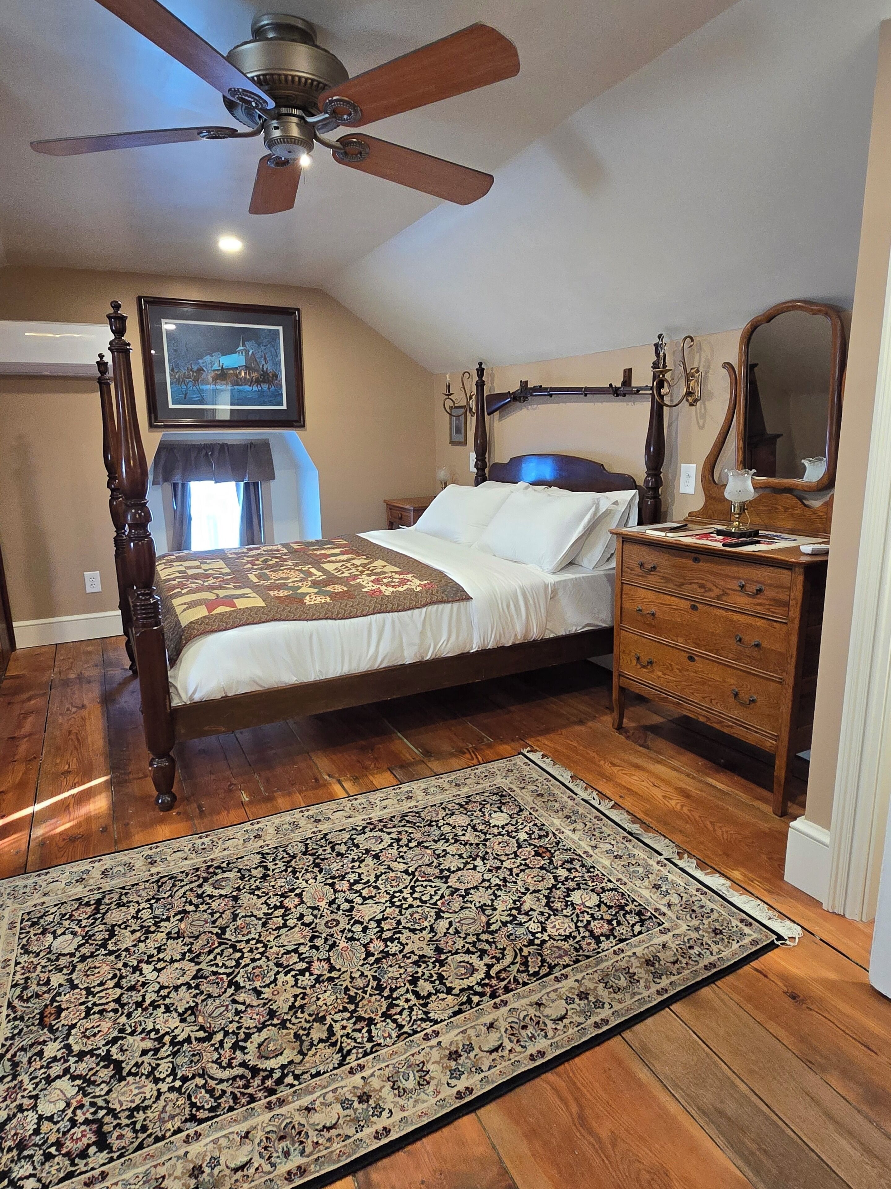 A bedroom with a four-poster bed, a wooden dresser, a ceiling fan, and a large rug.