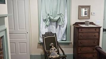 A room with a wooden floor, a dresser, a chair with a doll, a window with curtains, and a framed mirror on the wall.