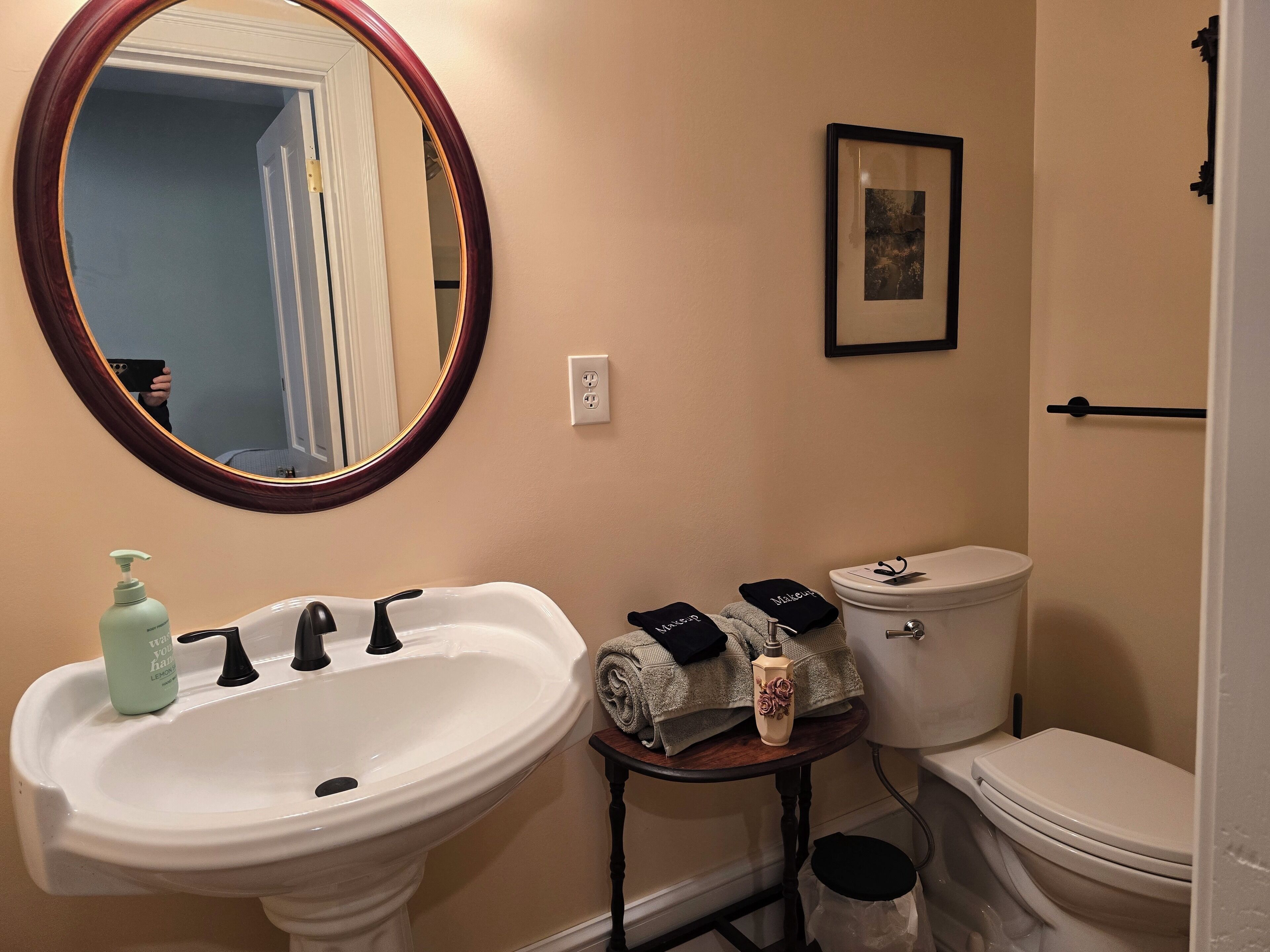 A bathroom with a white sink, a mirror, a toilet, a small table with towels, and a framed picture on the wall.