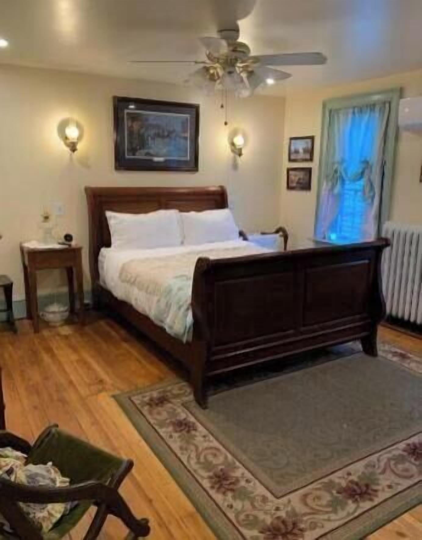 A bedroom with a bed, a ceiling fan, wall sconces, a framed picture, and a window with curtains.