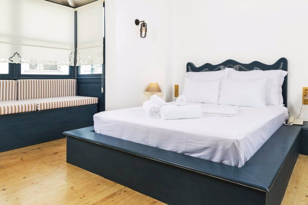 A bed with white linens, a bench with striped cushions, a wall-mounted lamp, and a window with lace curtains.