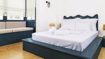 A bed with white linens, a bench with striped cushions, a wall-mounted lamp, and a window with lace curtains.