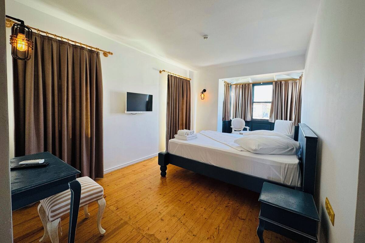 A bedroom with a bed, a desk, a chair, a TV, and a window with curtains.