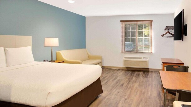 A hotel room with a bed, a desk, a chair, a lamp, a TV, and a window with blinds. - Woodspring Suites Charlotte Shelby (Shelby)