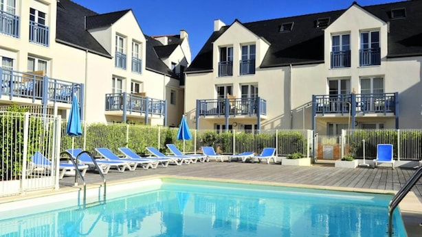 Una piscina con sillones de descanso y sombrillas, rodeada de edificios de apartamentos con balcones.