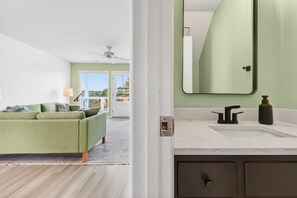 Una habitación con un sofá verde, un ventilador de techo blanco y un baño con un espejo grande y encimera de mármol.