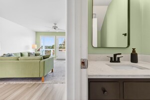 Una habitación con un sofá verde, un ventilador de techo blanco y un baño con un espejo grande y encimera de mármol.