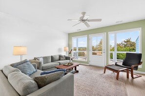 Ein Wohnzimmer mit Sofa, Couchtisch und Sessel. Es gibt einen Deckenventilator, eine Lampe und einen Blick durch große Glasfenster.