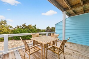 Una terraza de madera con una mesa y sillas, una barandilla blanca y un edificio azul al fondo.