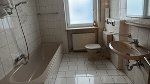 A bathroom with a bathtub, toilet, and sink, all with white fixtures and tiled walls. - Beautiful 3 room apartment on the first floor with terrace and garden area (Deggendorf)