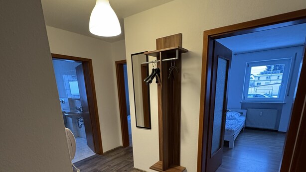A hallway with wooden flooring, a hanging light fixture, a mirror, and a door leading to a bedroom with a window. - Beautiful 3 room apartment on the first floor with terrace and garden area (Deggendorf)
