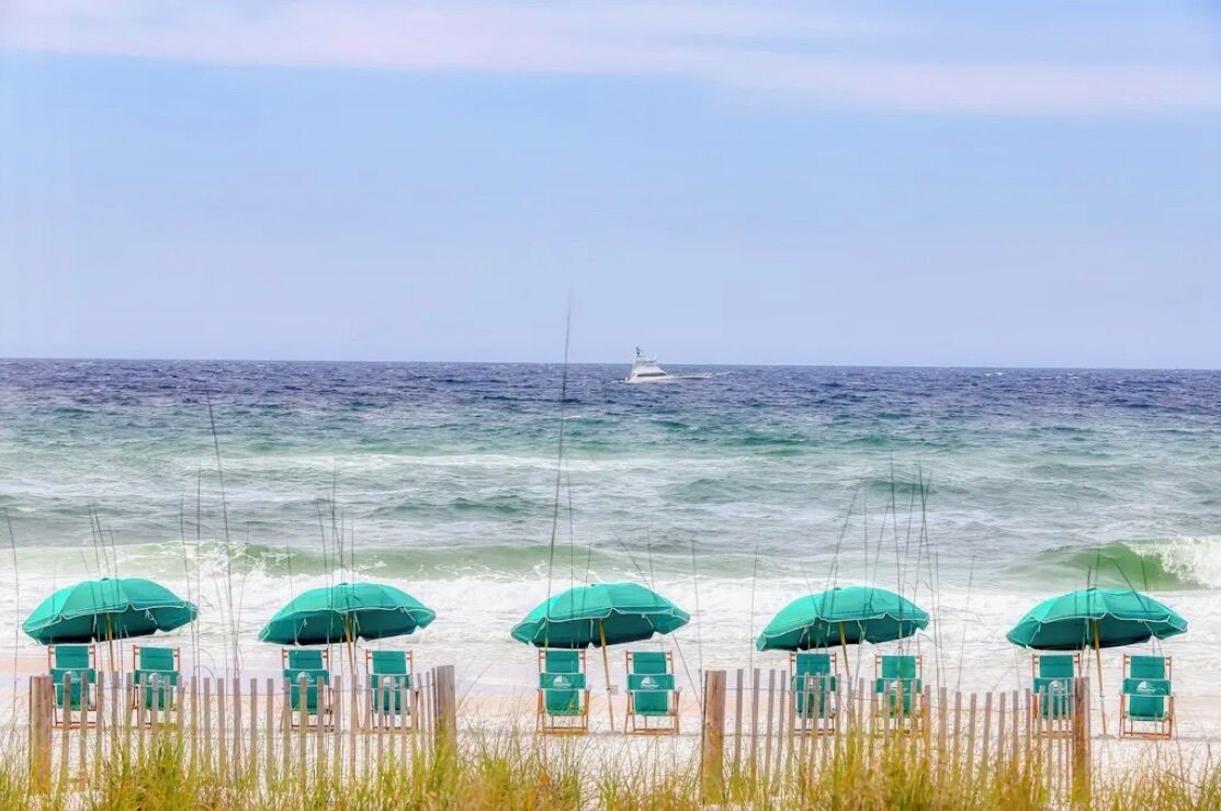 A beach with rows of green umbrellas and chairs, a wooden fence, and a boat in the water.