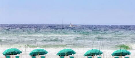 A beach with rows of green umbrellas and chairs, a wooden fence, and a boat in the water.