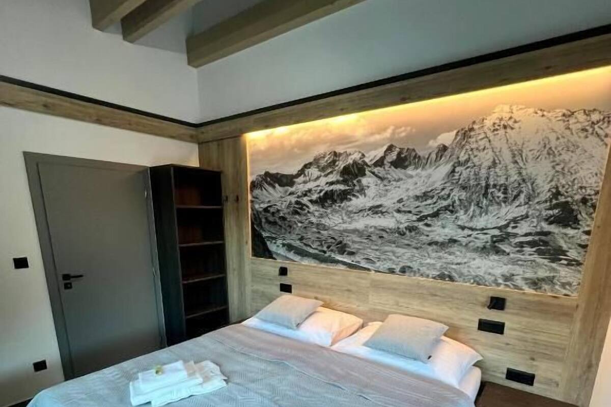 Une chambre à coucher avec un lit, des oreillers, une tête de lit ornée d’un tableau représentant une montagne, une armoire et un banc.