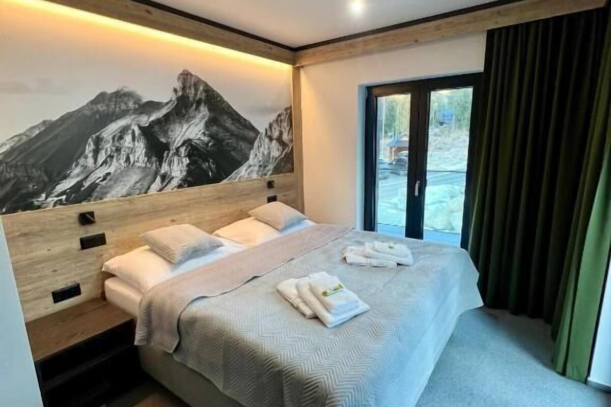 Une chambre d’hôtel moderne avec un lit, une fresque représentant une montagne, une fenêtre avec des rideaux et un luminaire au plafond.