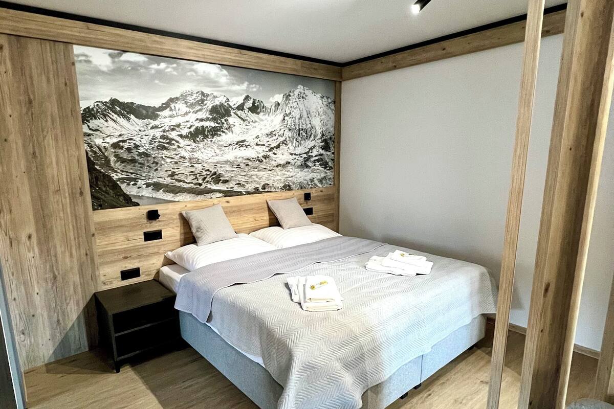 Une chambre à coucher moderne avec un lit, une tête de lit en bois et une grande fresque murale représentant un paysage de montagne.