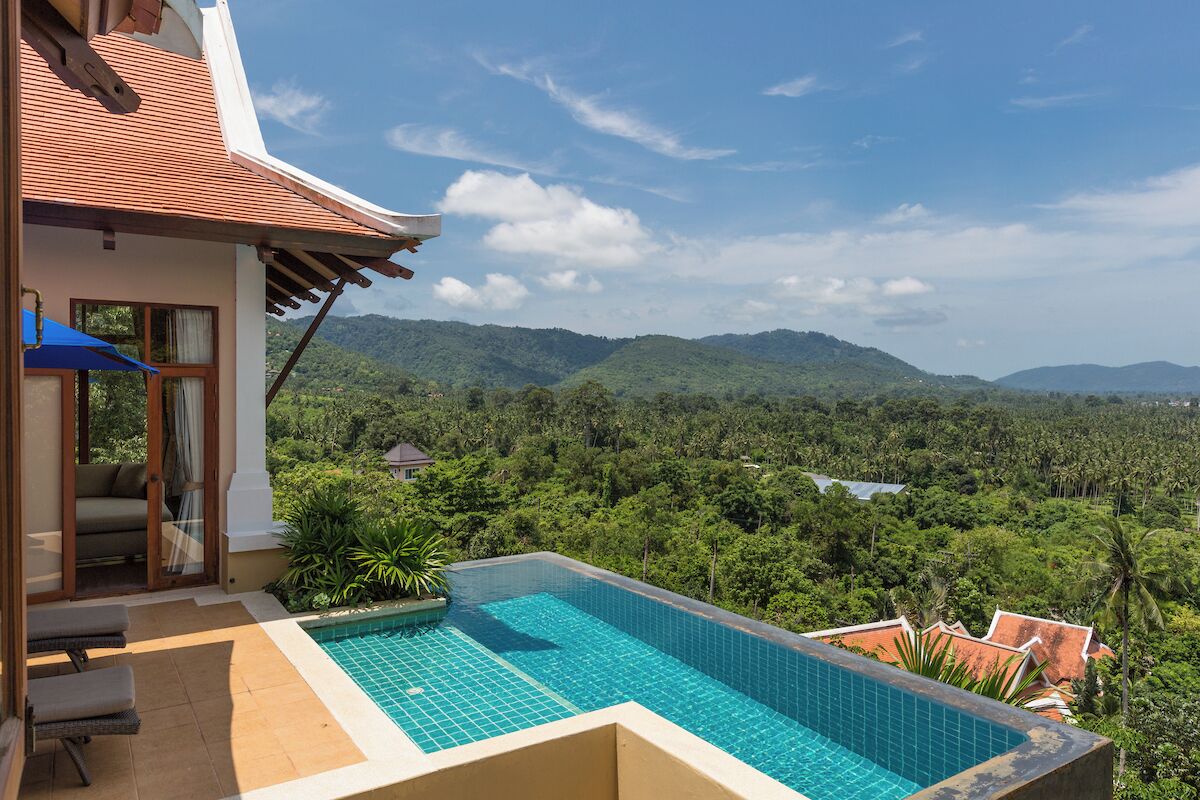 Une piscine offrant une vue sur un paysage luxuriant, un espace de détente couvert et un bâtiment doté d’un toit en tuiles.