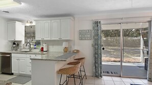Een keuken met witte kasten, een granieten aanrecht, een vast kookeiland met barkrukken, een accentmuur, en een schuifdeur naar een buitenruimte.