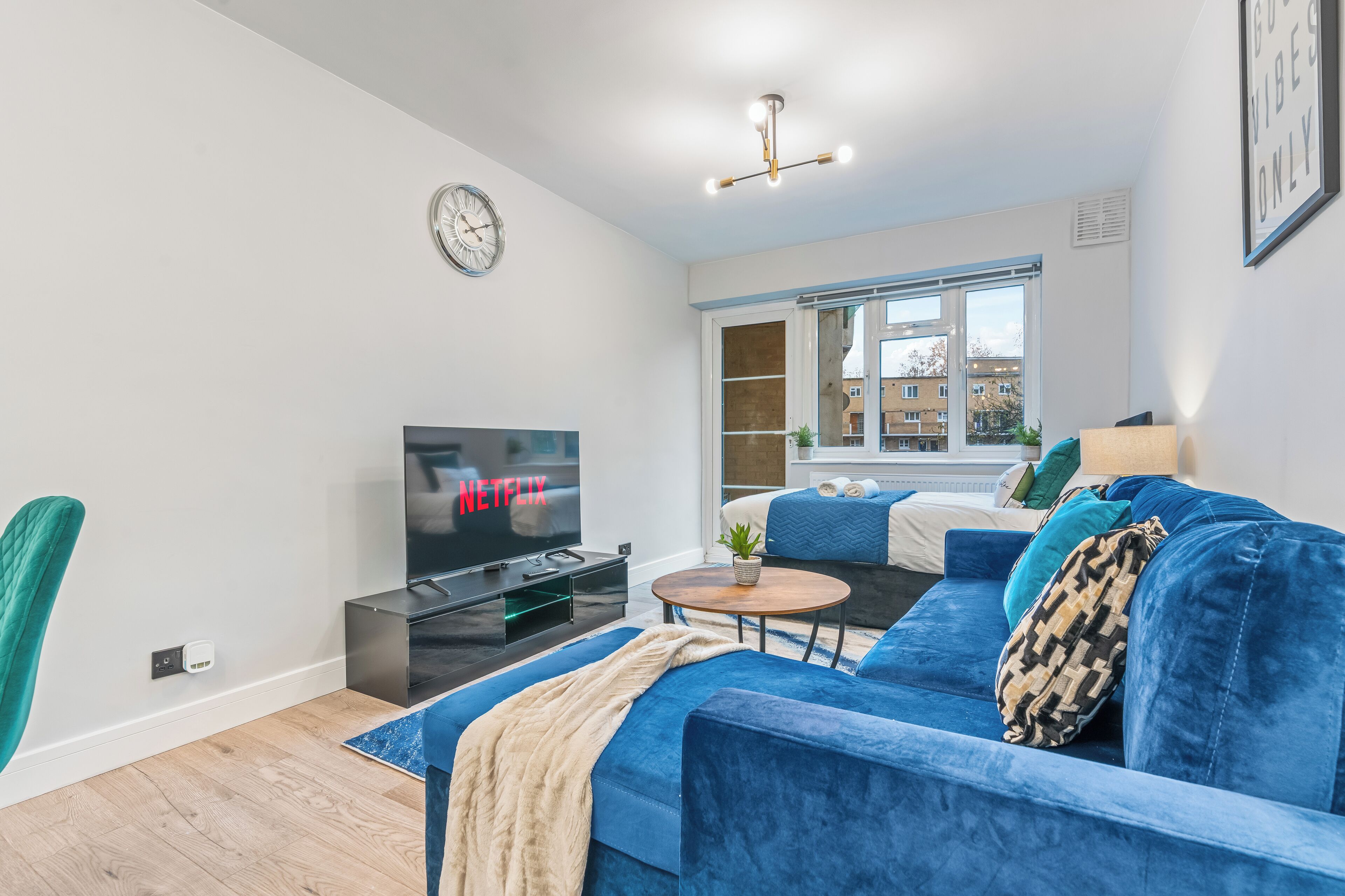 Un salon moderne avec un canapé bleu, un téléviseur, une horloge et une fenêtre donnant sur des immeubles.