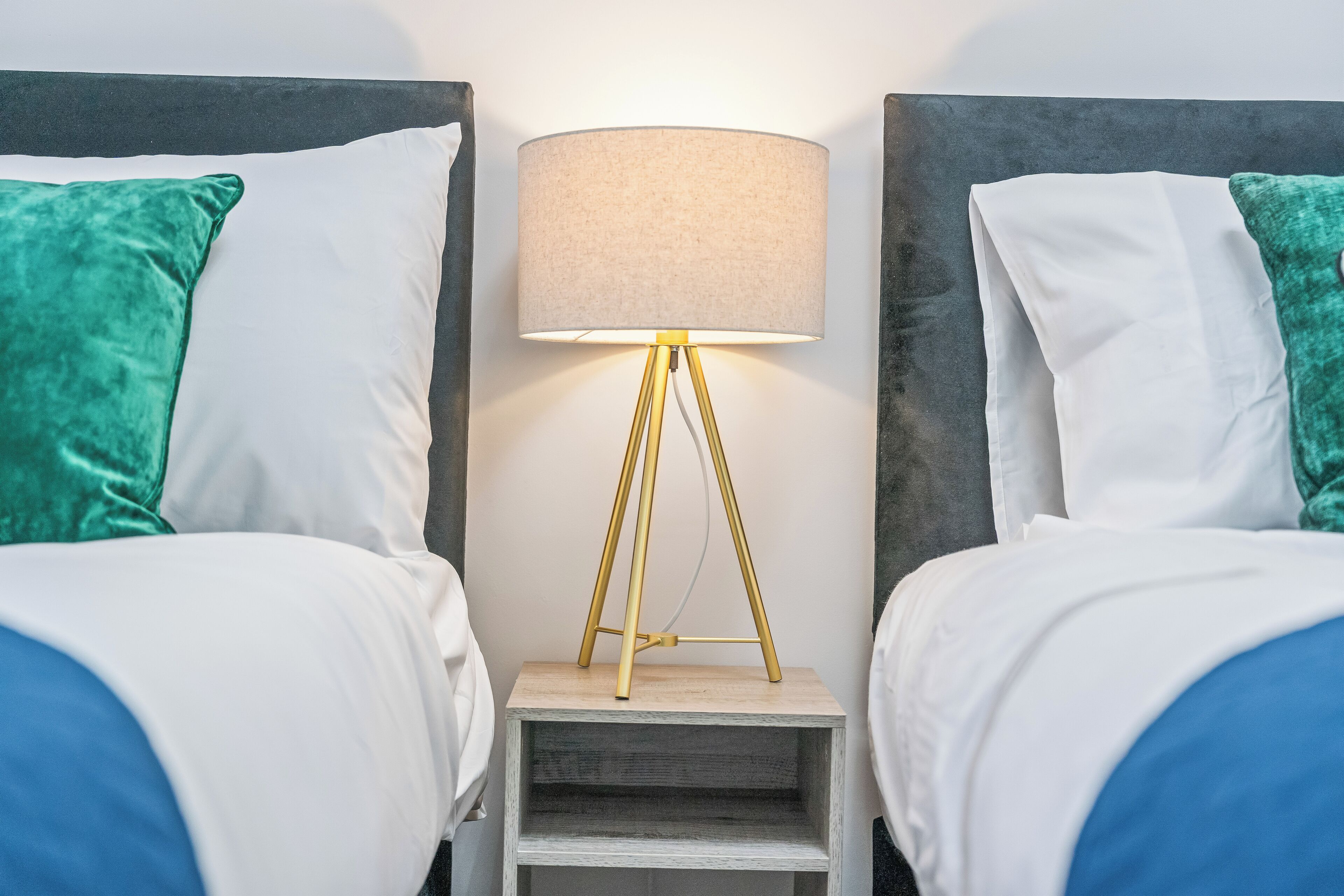 A hotel room with two beds, each with a white and green pillow, a bedside table with a lamp, and a small shelf.