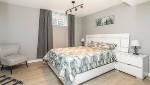 A bedroom with a bed, a chair, a bedside table with lamps, a window with curtains, and a painting on the wall.