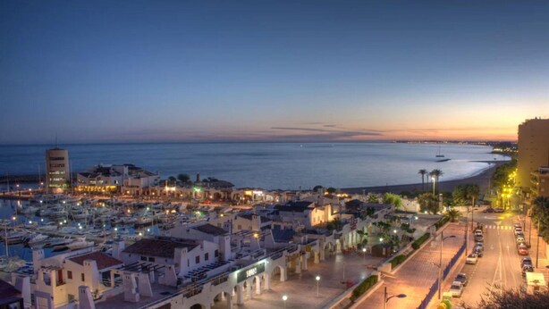 Un paisaje urbano costero al atardecer, con un puerto deportivo, edificios y una carretera que conduce a la playa.