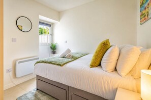 Ein Schlafzimmer mit einem Bett, Kissen, einer Nachttischlampe, einem Fenster mit Blick, einem Heizkörper und einem Wandspiegel.