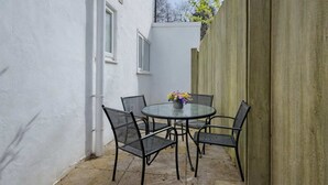 Une petite terrasse extérieure avec une table ronde et quatre chaises, un vase de fleurs et un mur blanc avec une fenêtre.