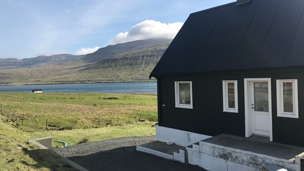A black house with white trim, a door, and windows, set against a backdrop of a lake and mountains.