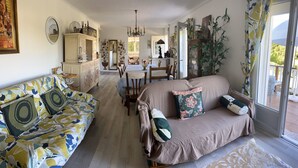 Una sala de estar con sofá, sillón y mesa de comedor con sillas. Hay un espejo grande, una araña y una planta en maceta.