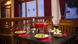 Une table à manger est dressée avec des assiettes, des verres et un pot rouge. De grandes fenêtres offrent une vue sur un paysage enneigé.